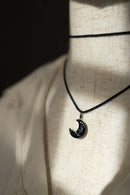 A black cord necklace with a crescent moon pendant featuring the word 'MEMENTO' engraved on it, displayed on a beige linen mannequin torso under dramatic shadow lighting.