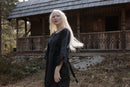 A woman with long platinum blonde hair stands outdoors in front of a rustic wooden cabin, wearing a loose black dress with blue patterned trim on the sleeves.