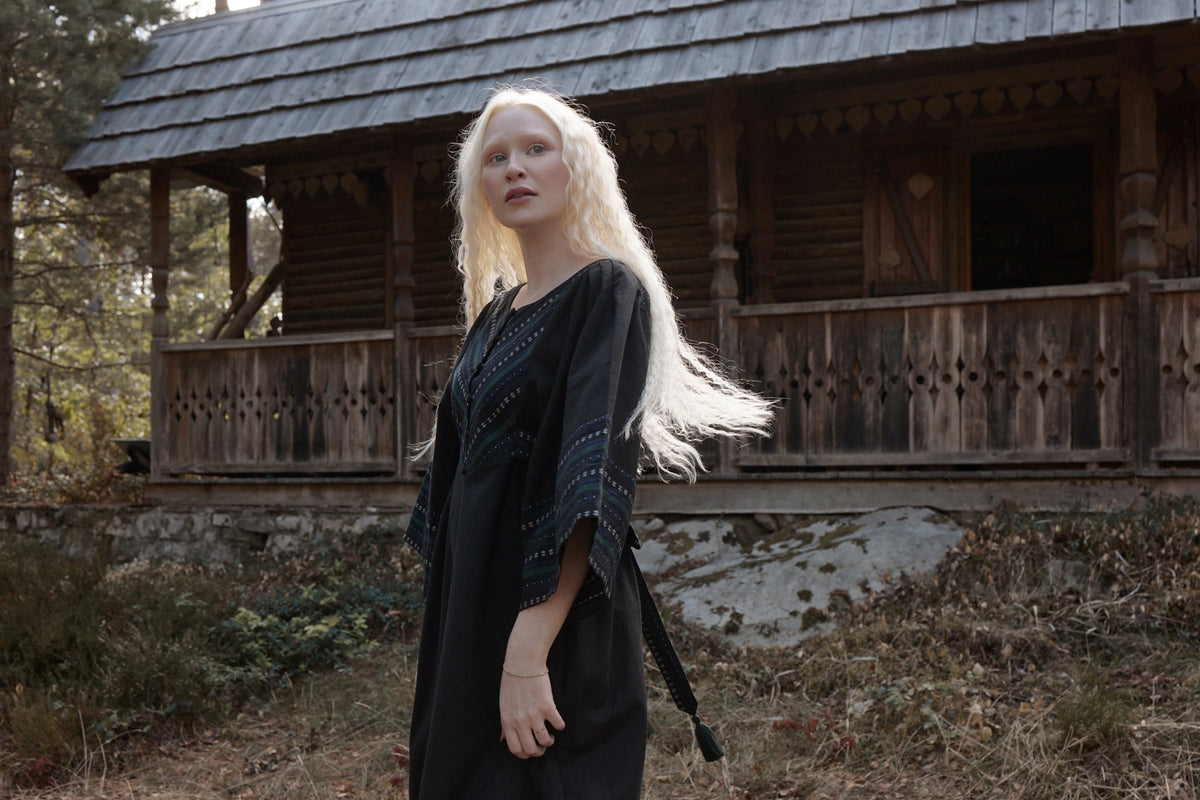 A woman with long platinum blonde hair stands outdoors in front of a rustic wooden cabin, wearing a loose black dress with blue patterned trim on the sleeves.