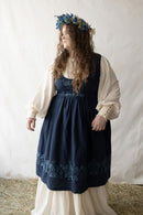 Woman plus size wearing a dark blue Bunad dress with floral embroidery and a white blouse, standing against a plain background.