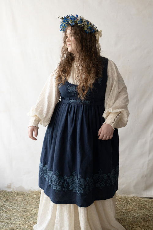 Woman plus size wearing a dark blue Bunad dress with floral embroidery and a white blouse, standing against a plain background.