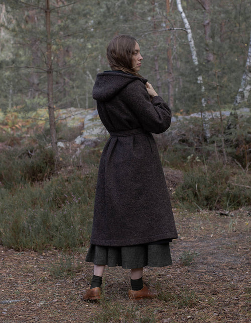 A woman stands outdoors in a wooded setting wearing a long dark brown textured coat over a dark skirt, with brown shoes and black socks.