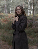 A woman wearing a dark charcoal gray wool-blend coat stands in a wooded outdoor setting, holding the collar of the coat closed.