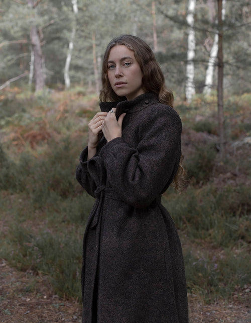 A woman wearing a dark charcoal gray wool-blend coat stands in a wooded outdoor setting, holding the collar of the coat closed.