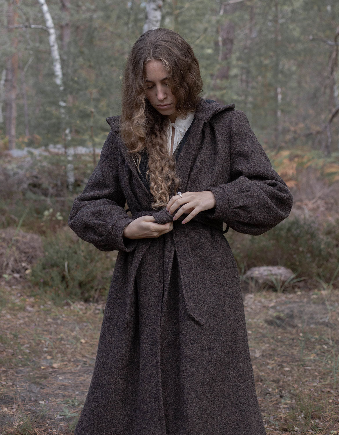 A woman wearing a dark brown textured wool coat with a hood and tie belt, standing in a wooded outdoor setting.
