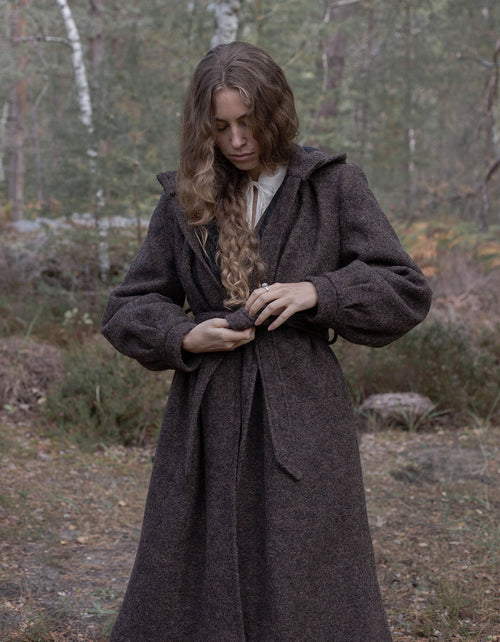 A woman wearing a dark brown textured wool coat with a hood and tie belt, standing in a wooded outdoor setting.