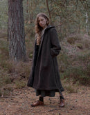 A woman stands in a wooded outdoor setting wearing a long dark charcoal-brown wool coat over a dark dress, with brown boots, trees and forest floor visible in the background.
