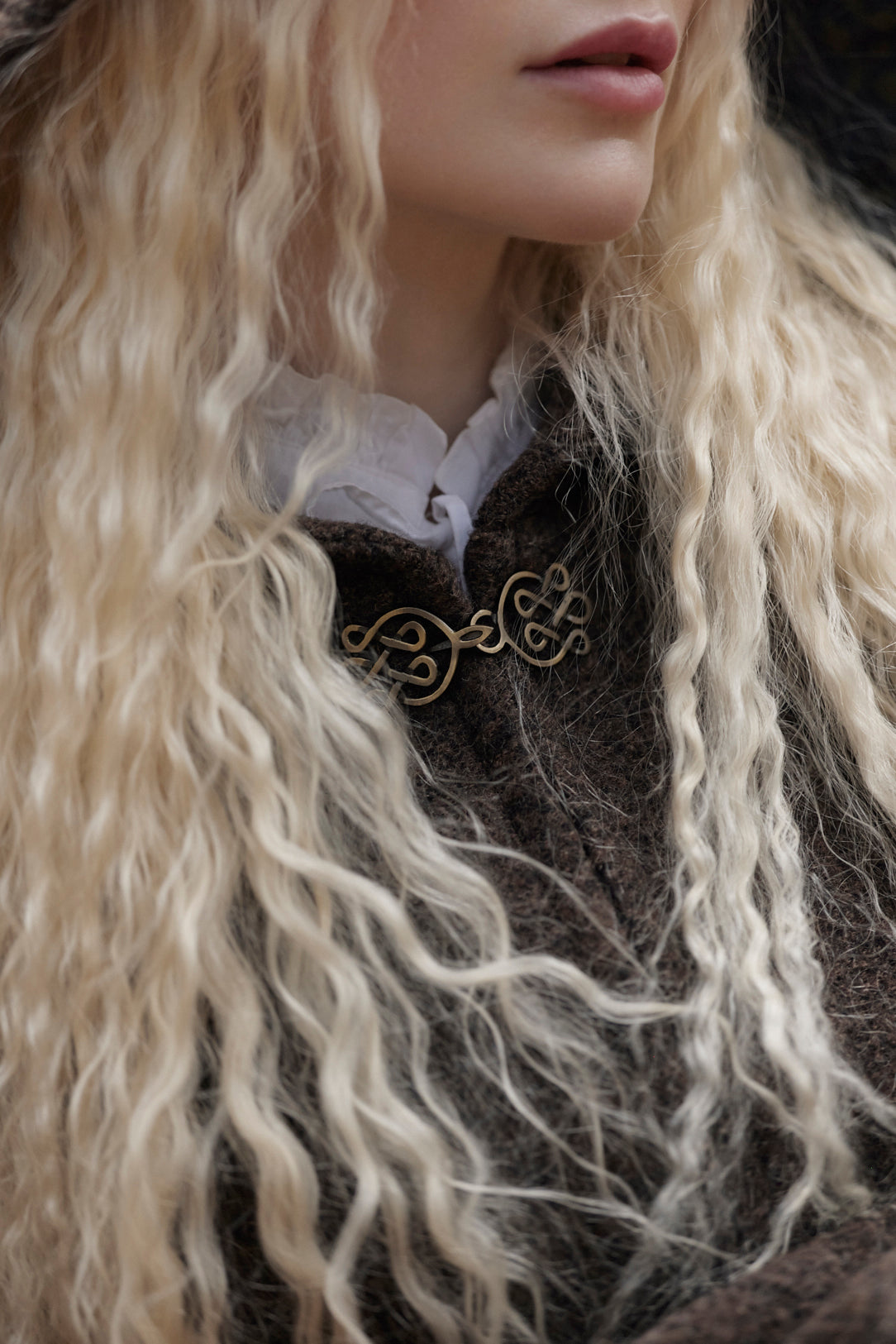 Close-up of a person with long, wavy platinum blonde hair wearing a dark brown textured coat over a white ruffled collar, with a gold-toned decorative brooch at the chest.