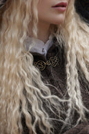 Close-up of a person with long, wavy platinum blonde hair wearing a dark brown textured coat over a white ruffled collar, with a gold-toned decorative brooch at the chest.