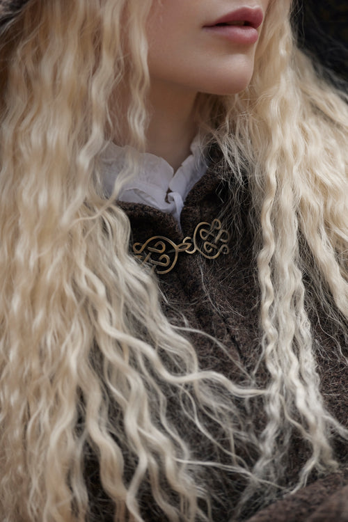 Close-up of a person with long, wavy platinum blonde hair wearing a dark brown textured coat over a white ruffled collar, with a gold-toned decorative brooch at the chest.