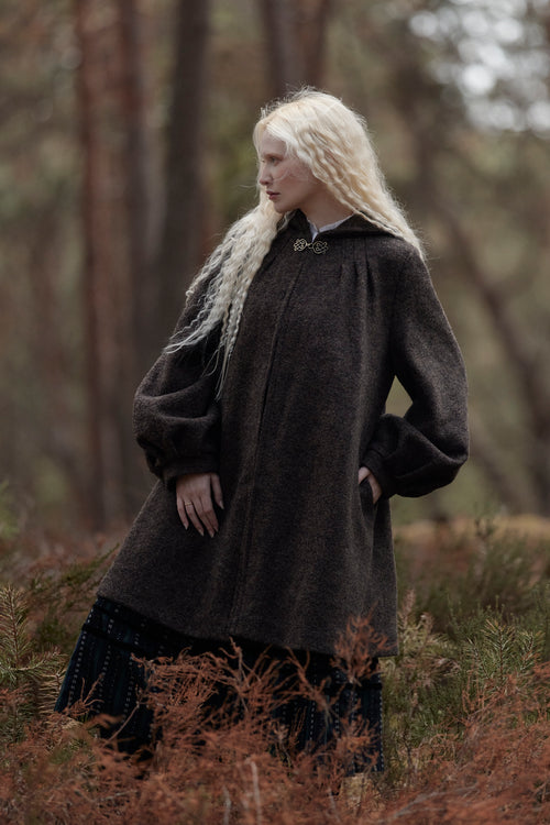 A woman with long platinum blonde hair stands in a forest clearing wearing a dark brown textured wool coat over a dark dress.
