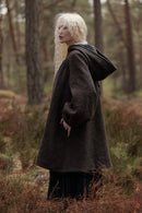 A woman with long platinum blonde hair stands in a forest wearing a dark brown textured hooded coat over a black dress.