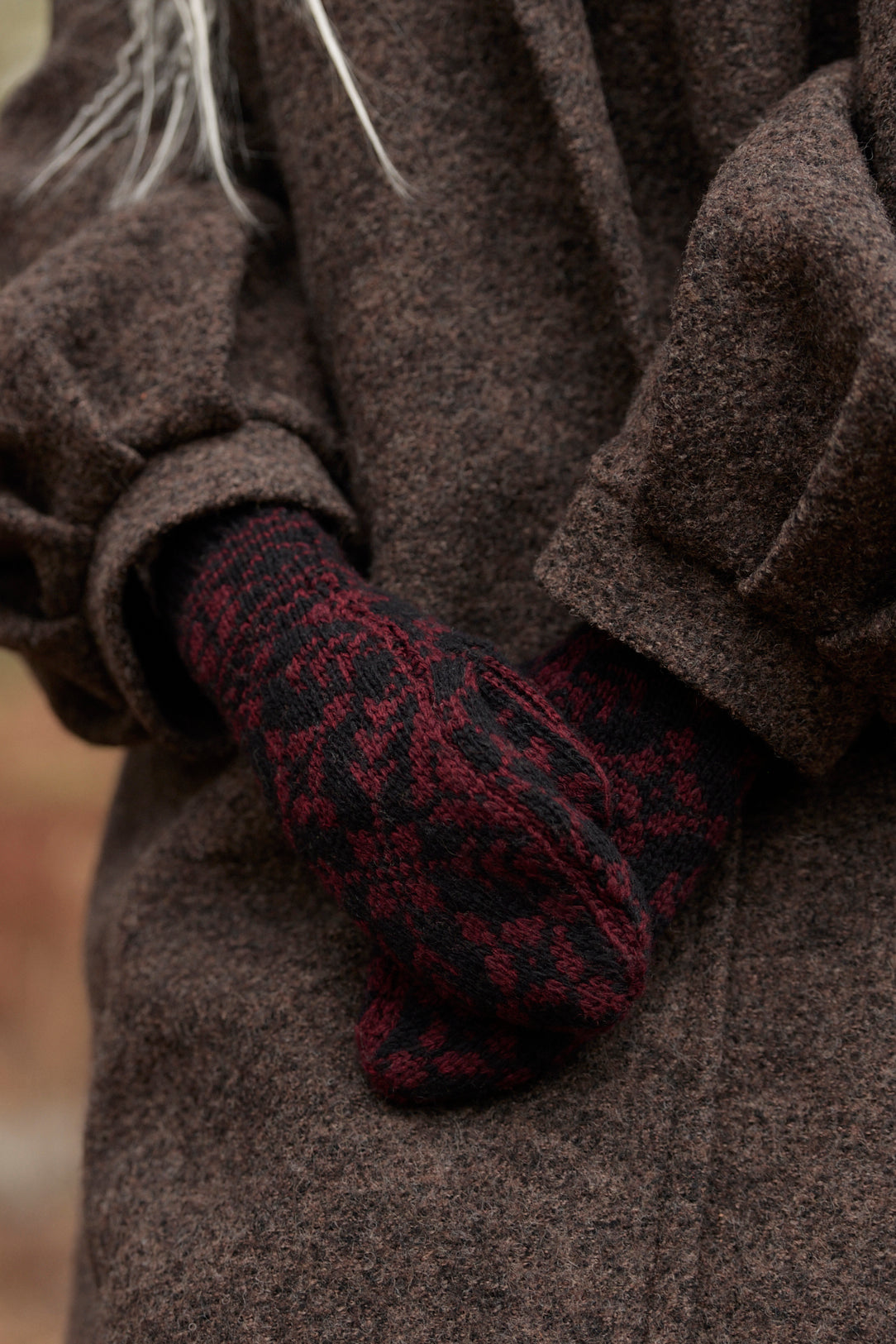 A close-up of a person wearing a dark brown textured coat and a dark red and black patterned knit glove, with hands tucked into the coat pocket.