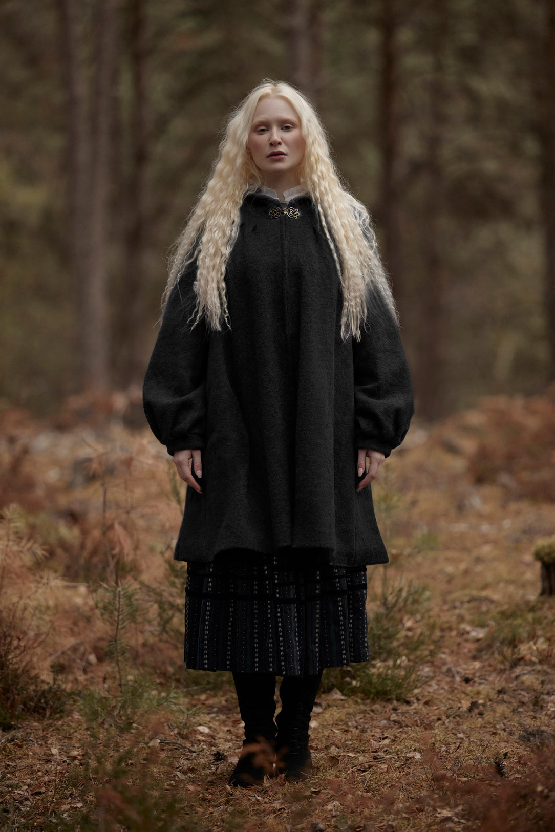 A woman with long platinum blonde hair stands in a forest wearing a dark charcoal gray woolen tunic with a high collar and a dark patterned skirt.