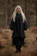 A woman with long platinum blonde hair stands in a forest wearing a dark charcoal gray woolen tunic with a high collar and a dark patterned skirt.