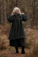 A woman with long platinum blonde hair stands in a forest wearing a dark charcoal gray woolen coat over a black pleated skirt and black boots.