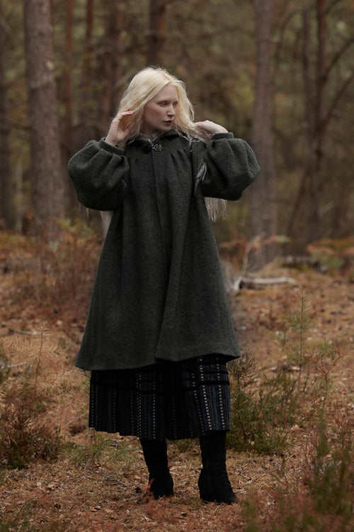 A woman with long platinum blonde hair stands in a forest wearing a dark charcoal gray woolen coat over a black pleated skirt and black boots.