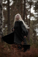 A woman with long platinum blonde hair stands in a forest wearing a voluminous black woolen coat with wide balloon sleeves. The forest background features tall trees and reddish-brown underbrush.
