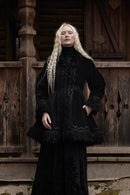 A woman with long blonde braids stands on a rustic wooden porch wearing a long black embroidered dress with dark fur trim at the collar, cuffs, and hem.