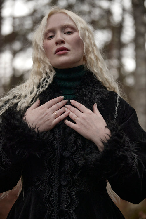 A woman with long, wavy platinum blonde hair wearing a black textured jacket with a dark green turtleneck, standing outdoors with her hands crossed over her chest.