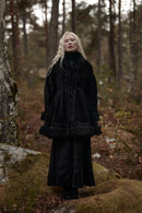 A woman with long blonde hair wearing a black long-sleeved dress with a ruffled collar and a matching black jacket with faux-fur trim, standing in a mossy forest.