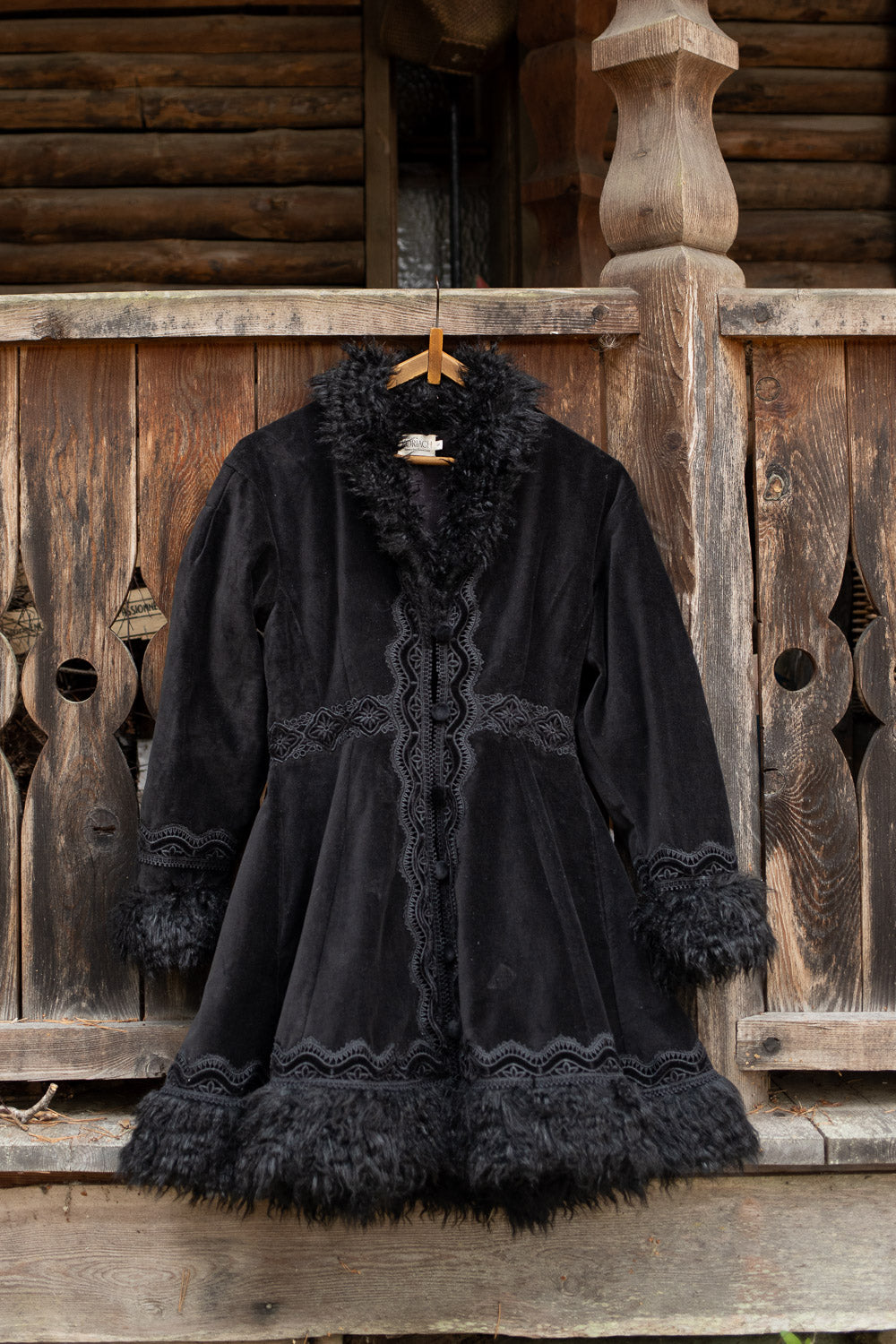 A black velvet coat with dark fur trim and lace detailing, hanging on a wooden hanger against a rustic wooden log cabin wall.