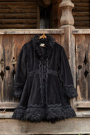 A black velvet coat with dark fur trim and lace detailing, hanging on a wooden hanger against a rustic wooden log cabin wall.
