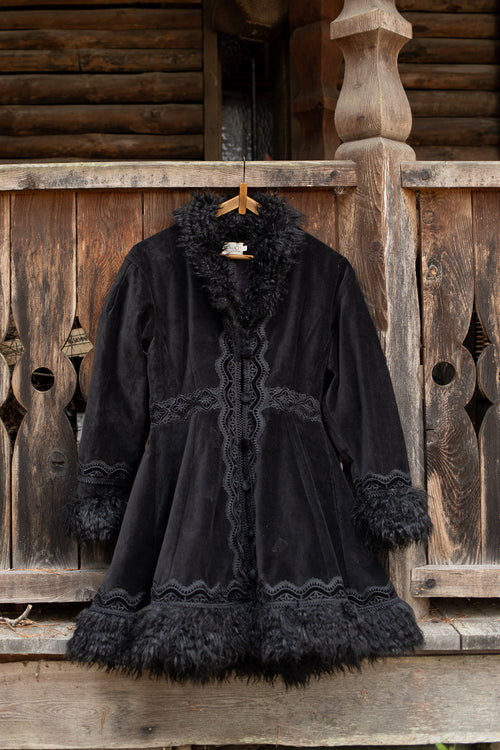 A black velvet coat with dark fur trim and lace detailing, hanging on a wooden hanger against a rustic wooden log cabin wall.