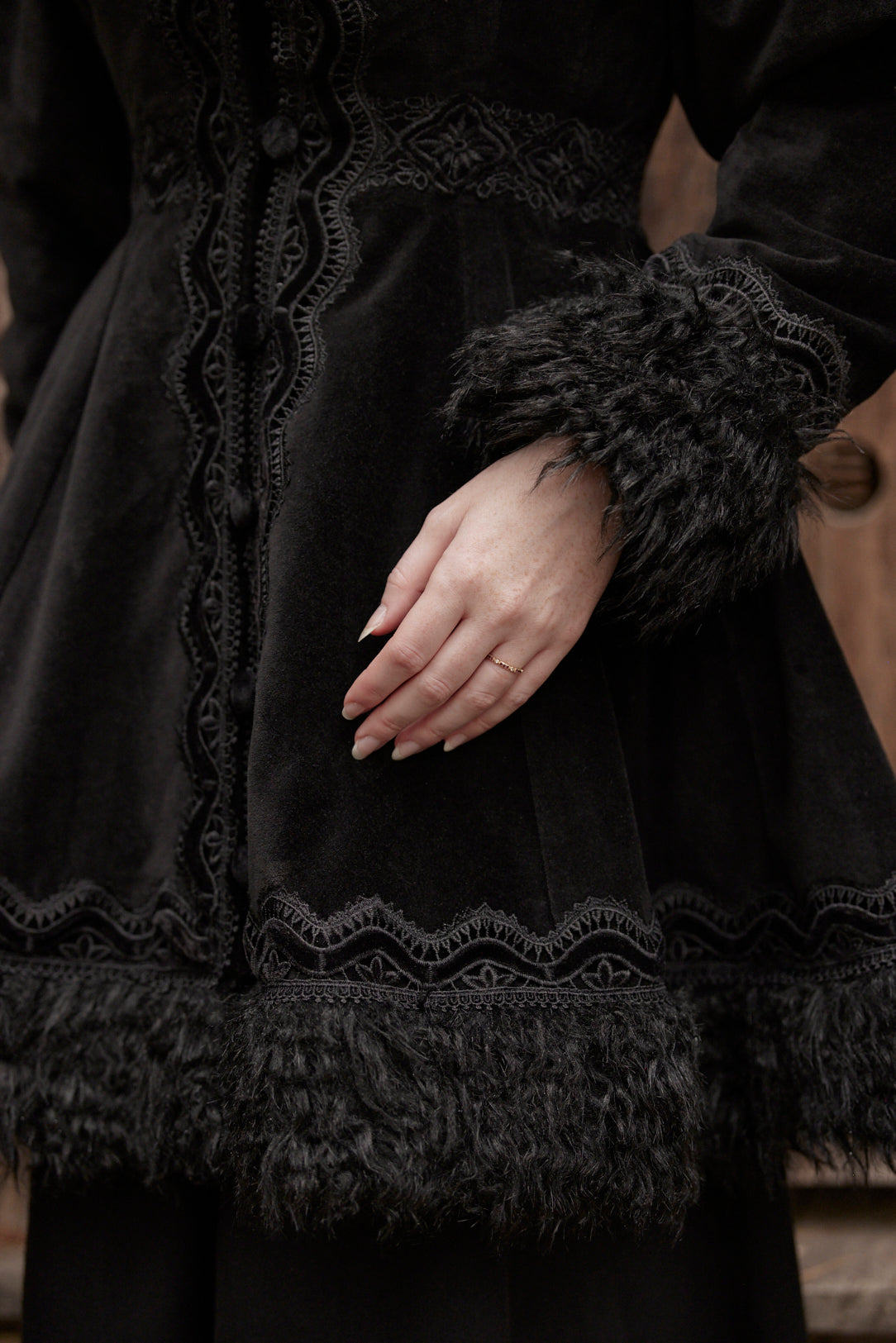 Close-up of a person wearing a black vintage-style dress with lace trim, textured fabric, and fluffy trim on the cuffs and hem, with one hand resting on the skirt.