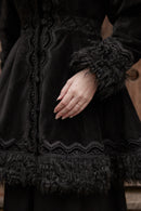 Close-up of a person wearing a black vintage-style dress with lace trim, textured fabric, and fluffy trim on the cuffs and hem, with one hand resting on the skirt.