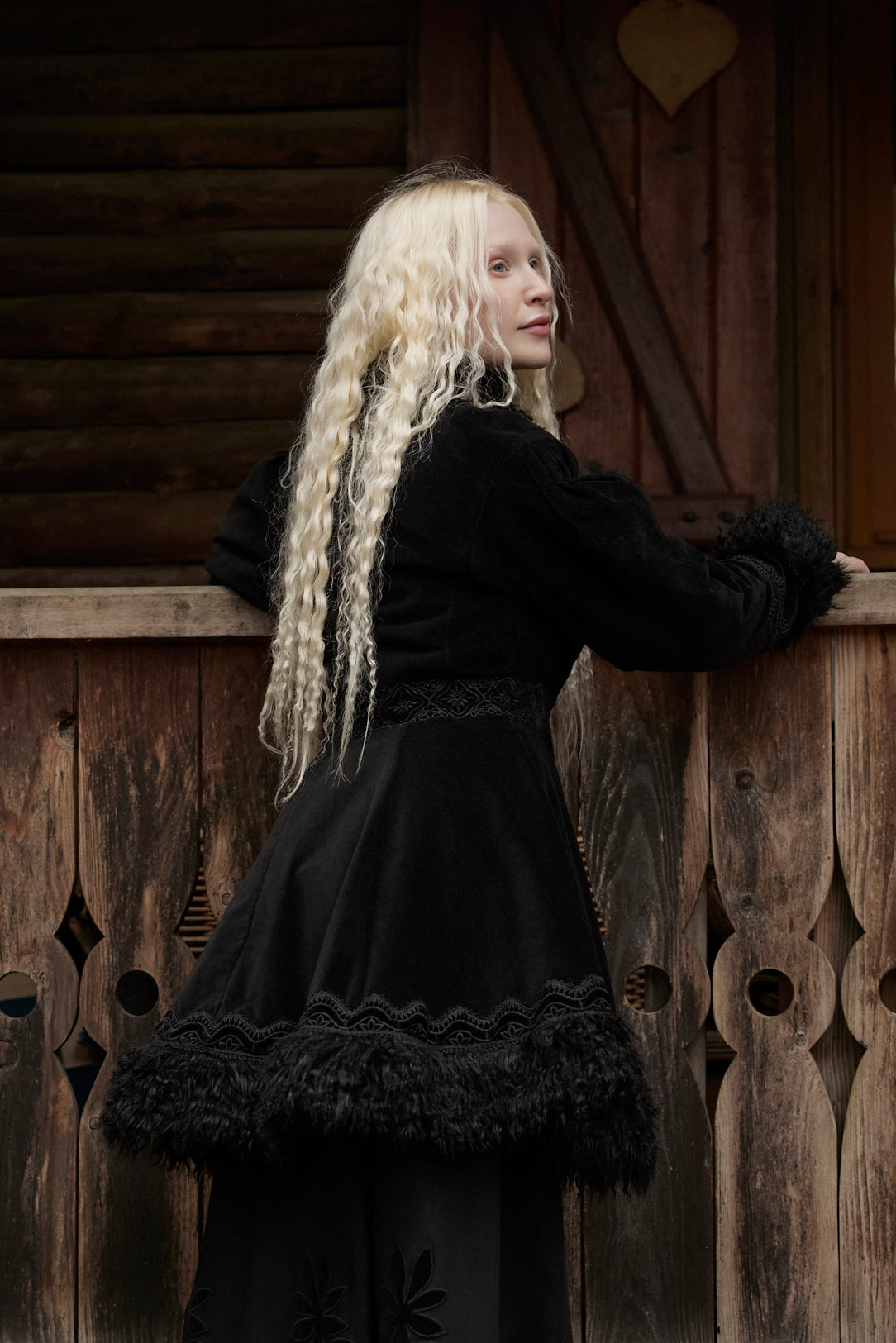 A woman with long, curly platinum blonde hair is shown from behind, wearing a black medieval-style dress with a ruffled tulle hem and dark feather trim, leaning against a rustic wooden fence.