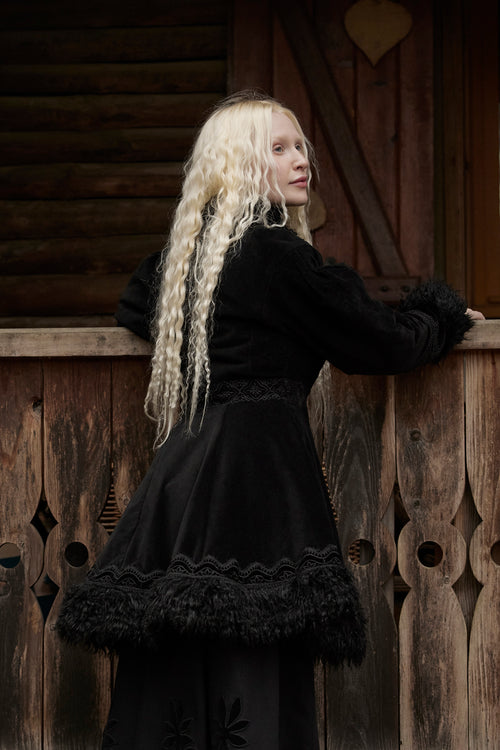 A woman with long, curly platinum blonde hair is shown from behind, wearing a black medieval-style dress with a ruffled tulle hem and dark feather trim, leaning against a rustic wooden fence.