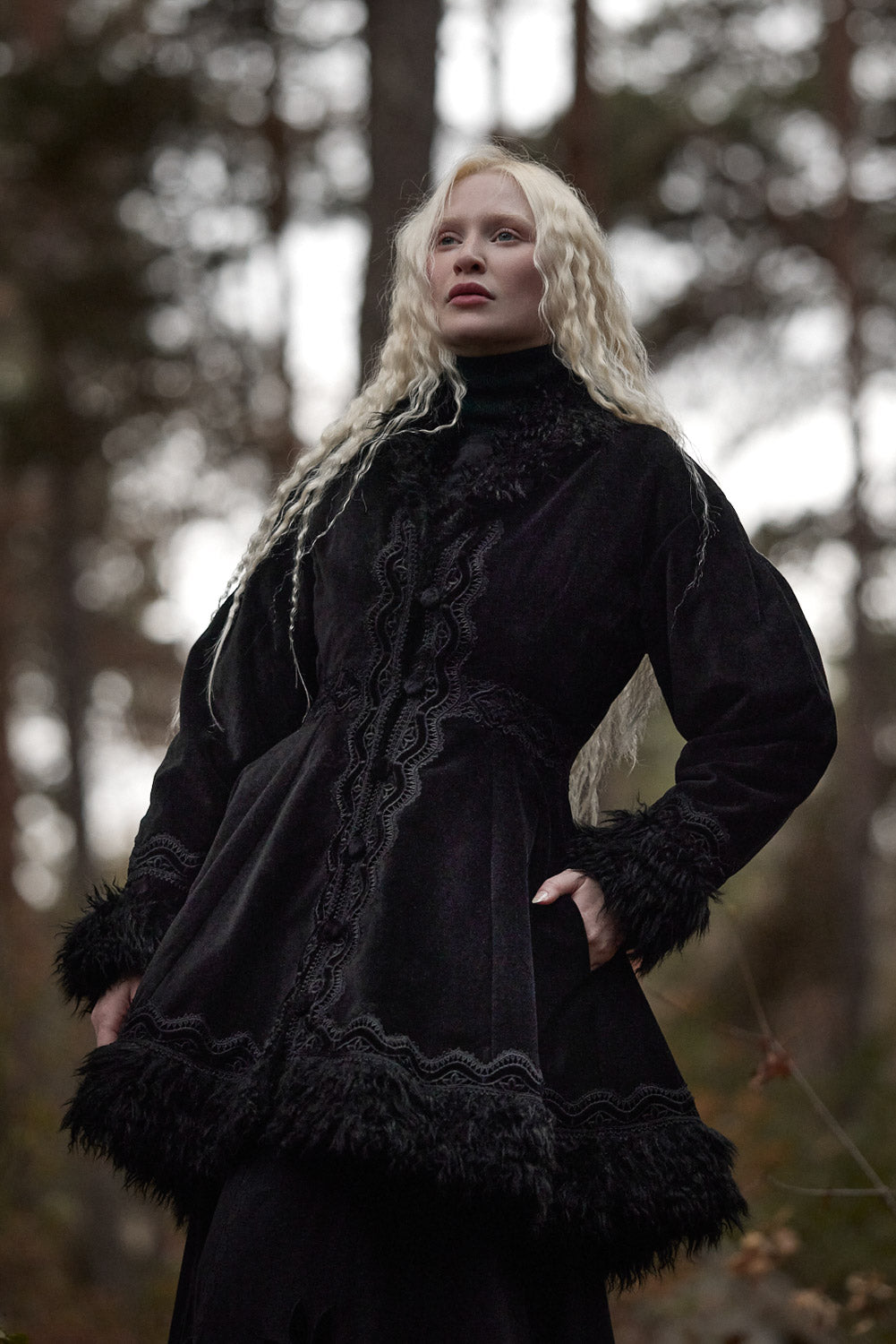 A woman with long platinum blonde hair wearing a black velvet gothic-style coat with lace trim and faux fur details, standing in a blurred forest setting.