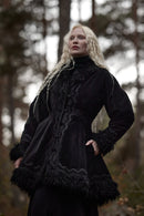 A woman with long platinum blonde hair wearing a black velvet gothic-style coat with lace trim and faux fur details, standing in a blurred forest setting.