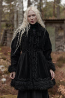 A woman with long platinum blonde hair stands outdoors in a forest-like setting wearing a voluminous black coat with elaborate lace trim, ruffles, and dark fringe details.