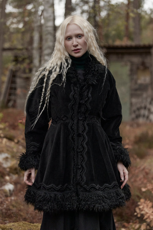 A woman with long platinum blonde hair stands outdoors in a forest-like setting wearing a voluminous black coat with elaborate lace trim, ruffles, and dark fringe details.