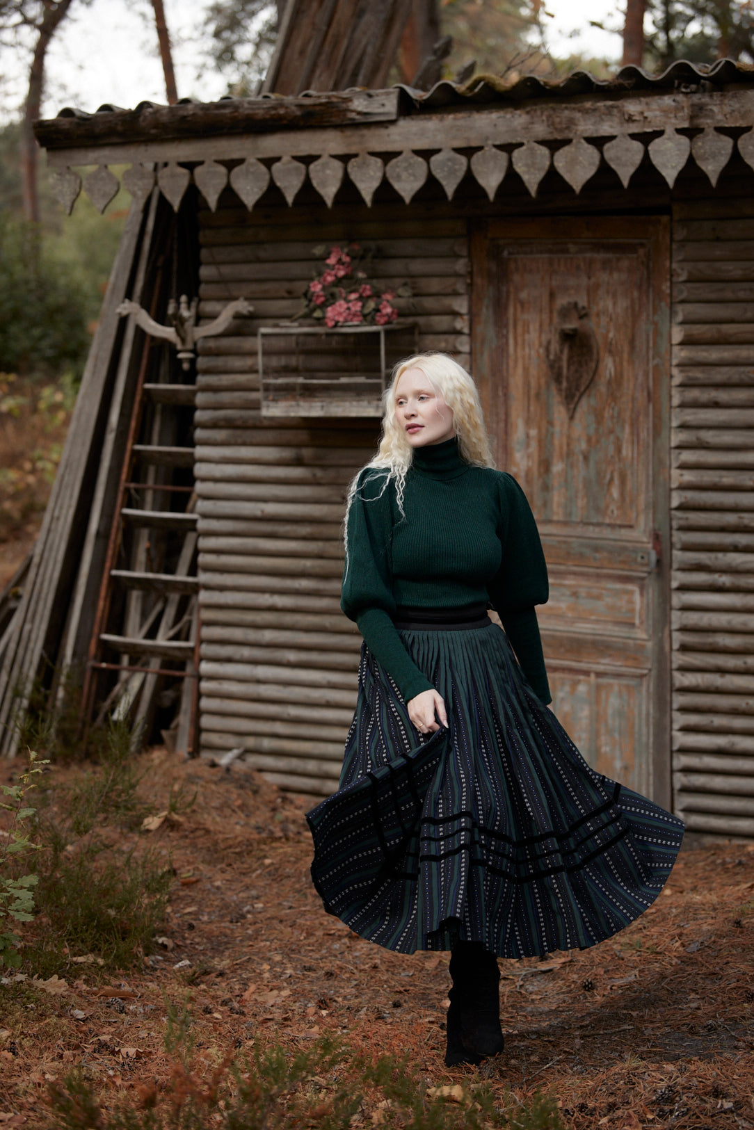 A woman with platinum blonde hair stands in front of a rustic wooden cabin, wearing a dark green turtleneck sweater and a dark pleated skirt with white vertical stripes.