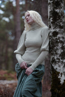 A woman with long blonde hair stands outdoors against a tree trunk in a forest, wearing a ribbed beige turtleneck sweater and a dark green skirt.