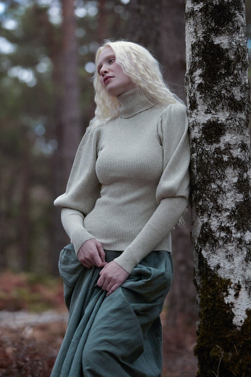 A woman with long blonde hair stands outdoors against a tree trunk in a forest, wearing a ribbed beige turtleneck sweater and a dark green skirt.