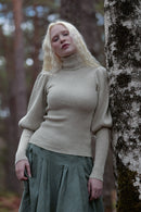 A woman with long blonde hair stands outdoors next to a tree trunk, wearing a ribbed beige turtleneck sweater and a dark green skirt.