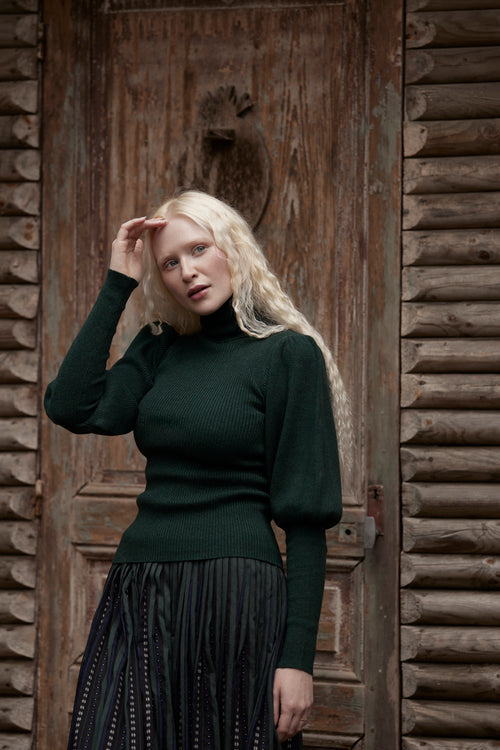 A blonde woman posing in front of a rustic wooden door, wearing a dark forest green ribbed turtleneck sweater and a dark pleated skirt.
