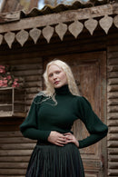 A blonde woman wearing a dark green ribbed turtleneck sweater and a dark pleated skirt stands in front of a rustic wooden cabin door.