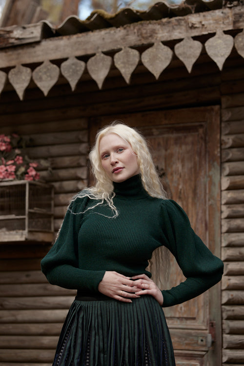 A blonde woman wearing a dark green ribbed turtleneck sweater and a dark pleated skirt stands in front of a rustic wooden cabin door.