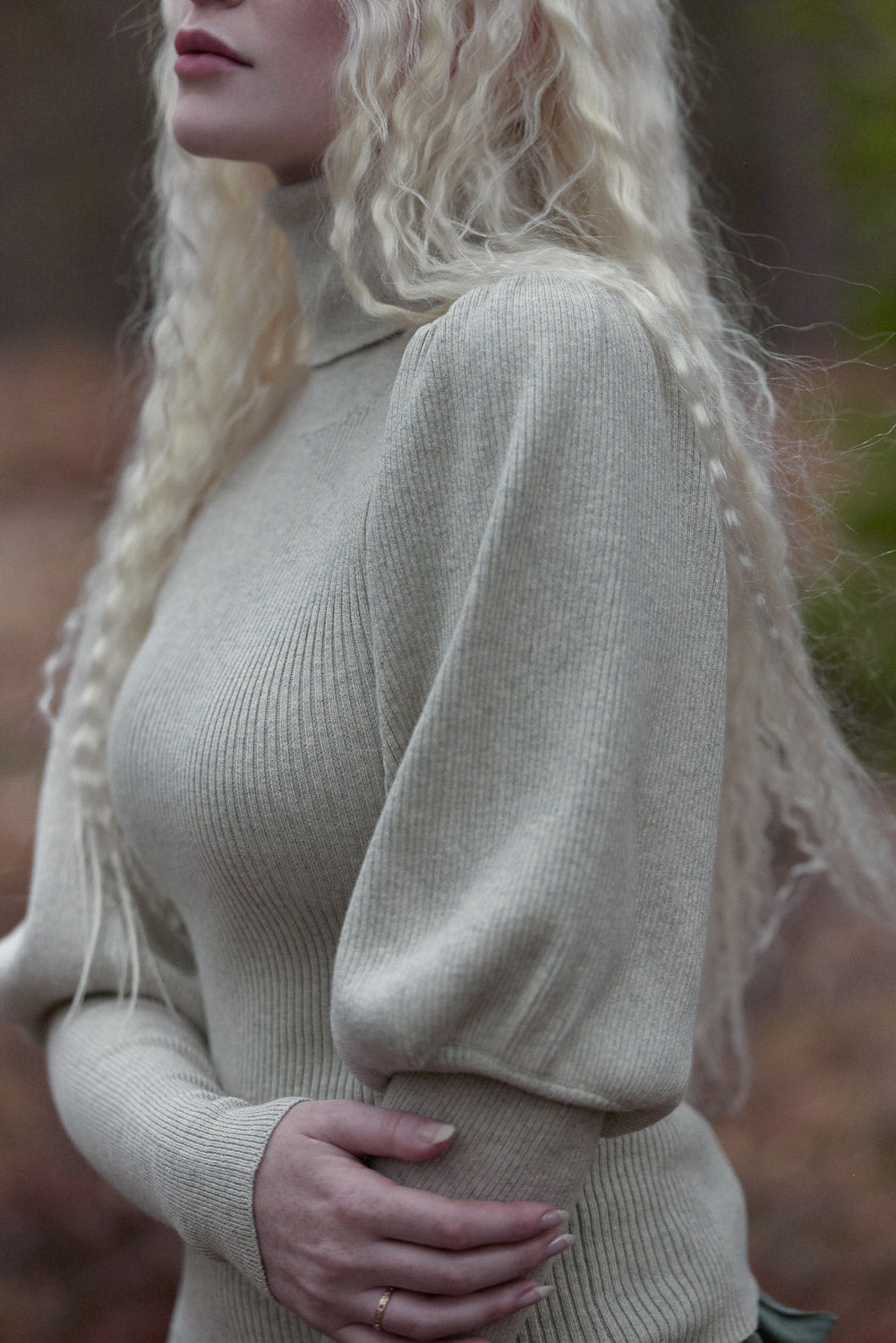 A close-up side view of a woman wearing a ribbed knit turtleneck sweater in a light oatmeal beige color, with long blonde hair and a blurred outdoor background.