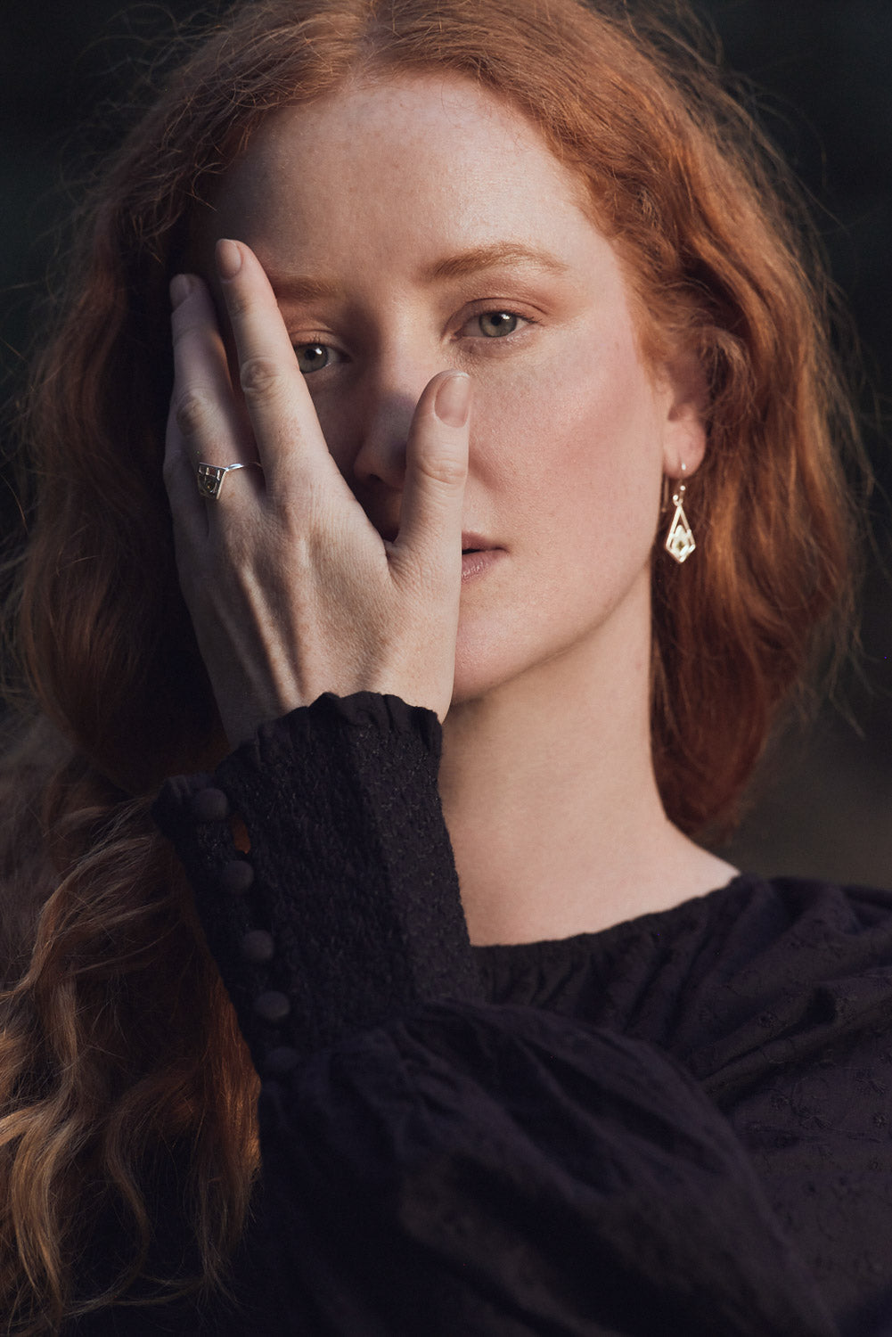 Woman with red hair wearing a black top and earrings, holding her hand to her face.