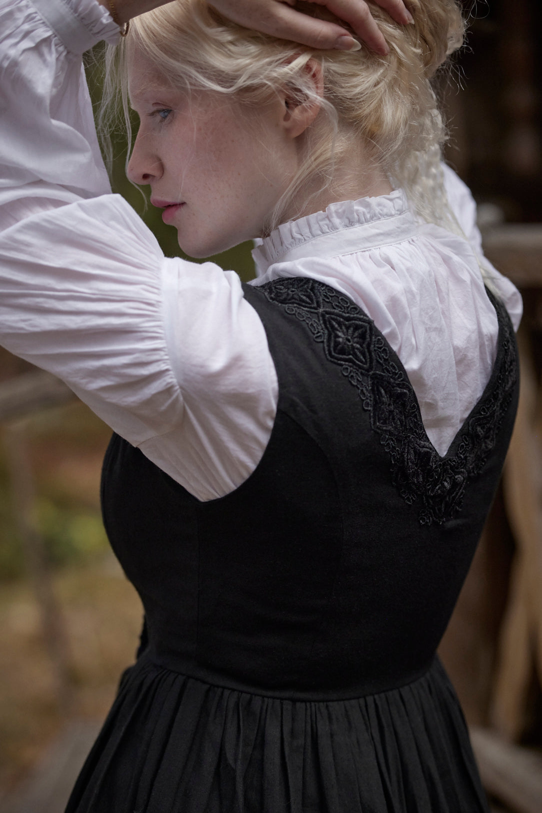 A woman wearing a black medieval-style dress with a white ruffled blouse, shown from the back in an outdoor setting.