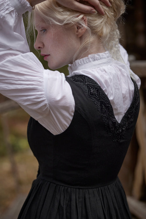 A woman wearing a black medieval-style dress with a white ruffled blouse, shown from the back in an outdoor setting.