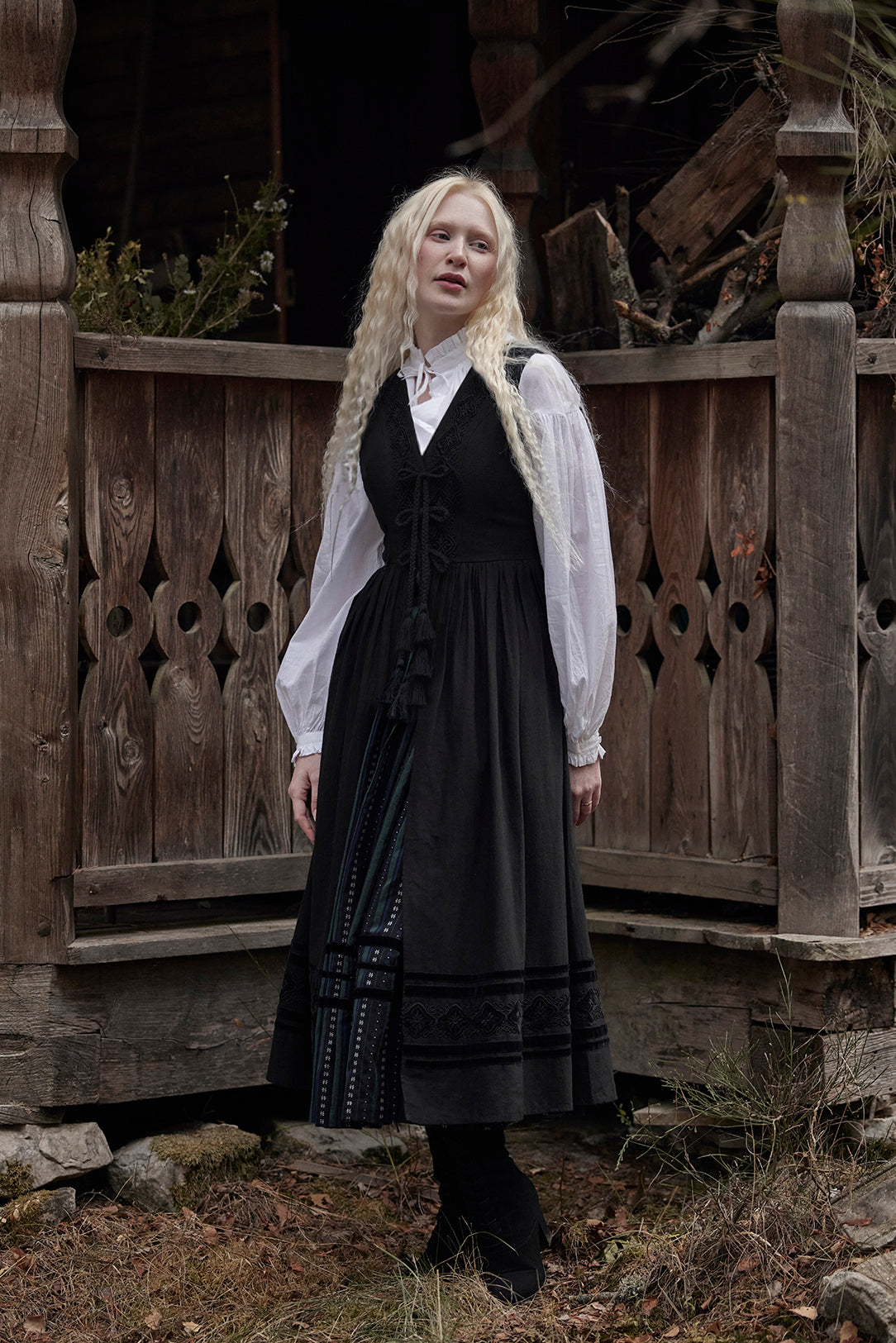 A woman with long blonde braids stands outdoors in front of a rustic wooden structure, wearing a black traditional-style dress with a white sheer long-sleeve blouse layered underneath.