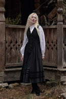 A woman with long blonde braids stands outdoors in front of a rustic wooden structure, wearing a black traditional-style dress with a white sheer long-sleeve blouse layered underneath.
