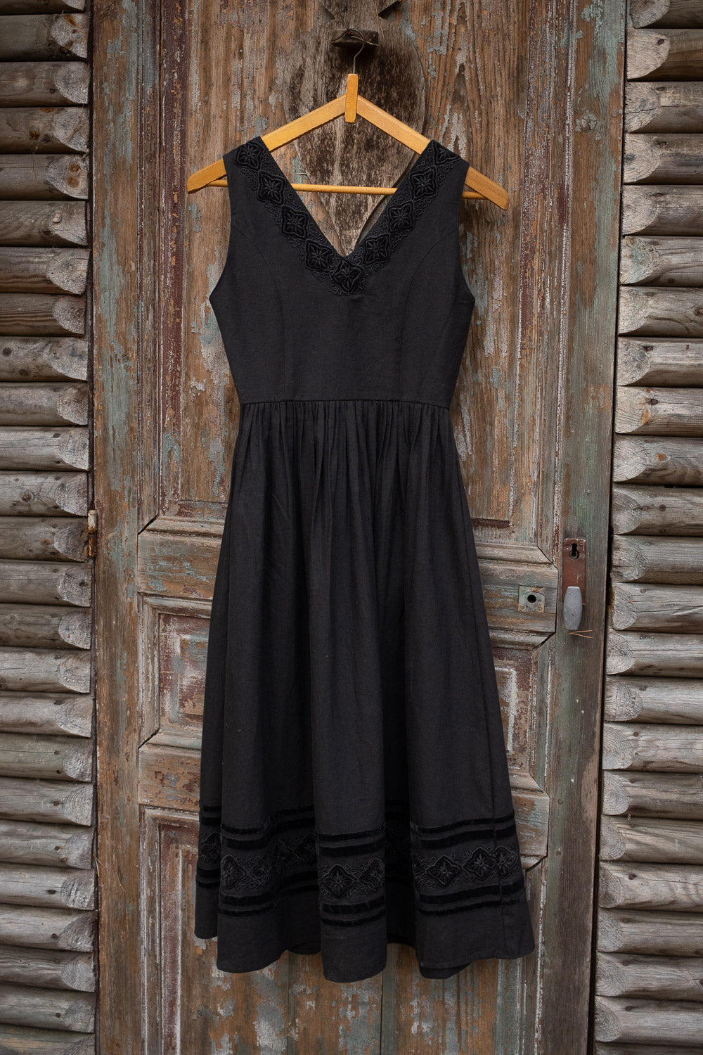 A sleeveless black midi dress with lace trim at the V-neckline and hem, hanging on a wooden hanger against a rustic weathered wooden door.